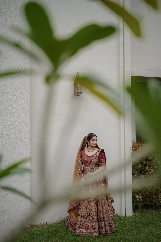 Bride in a maroon and gold embroidered lehenga standing against a white wall, framed by leaves
